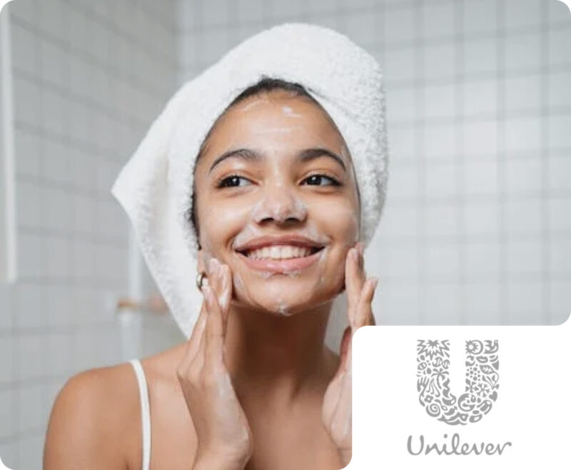 A smiling woman with a towel-wrapped head applying facial cleanser, with a Unilever brand badge. This illustrates Flipflow's capability in monitoring market presence, product performance, and consumer sentiment for global personal care and beauty brands.