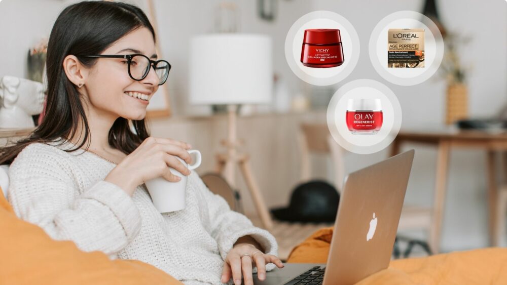 oung woman with glasses smiling on a sofa, working on a laptop and holding a white mug, with floating product images of Vichy Liftactiv, L’Oréal Age Perfect and Olay Regenerist face creams beside her.