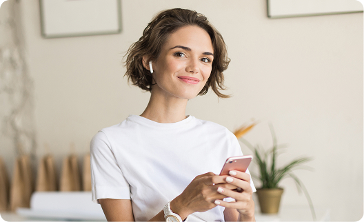 A smiling young woman wearing a white t-shirt and wireless earbuds holds a smartphone, looking directly at the camera in a brightly lit indoor setting.