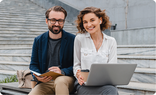 A happy male and female colleague smiling while sitting on urban steps. The man is holding a notebook, and the woman is working on a laptop with a coffee cup nearby, suggesting casual outdoor business collaboration.