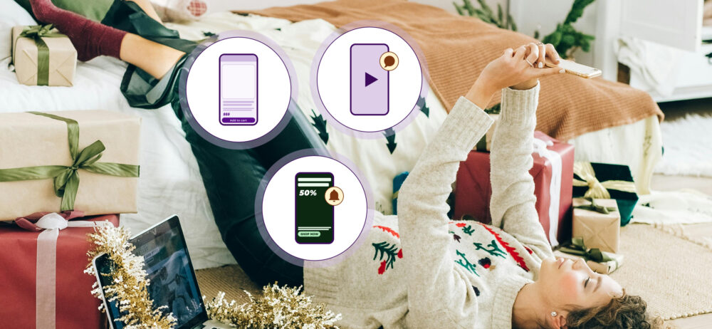 Escena Mobile Commerce Navidad 2025: persona recostada en una cama entre regalos usando el teléfono; iconos de “Add to cart”, video y oferta “50% / Shop now”.