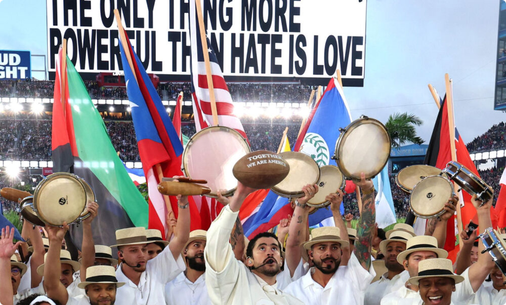 Percussionists in a Super Bowl-style stadium lifting drums and flags in front of a giant screen during a show.