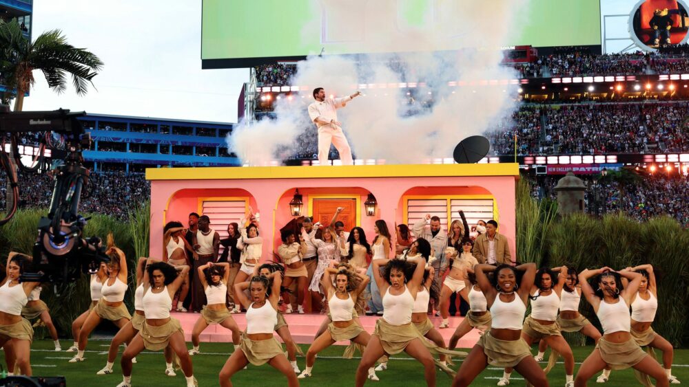 Performance in a Super Bowl-style stadium with a pink house as a stage, smoke, and a large dance troupe in light-coloured outfits.