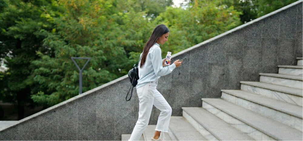 Mujer subiendo escaleras mientras mira el móvil y lleva un yogur bebible on-the-go en la mano.