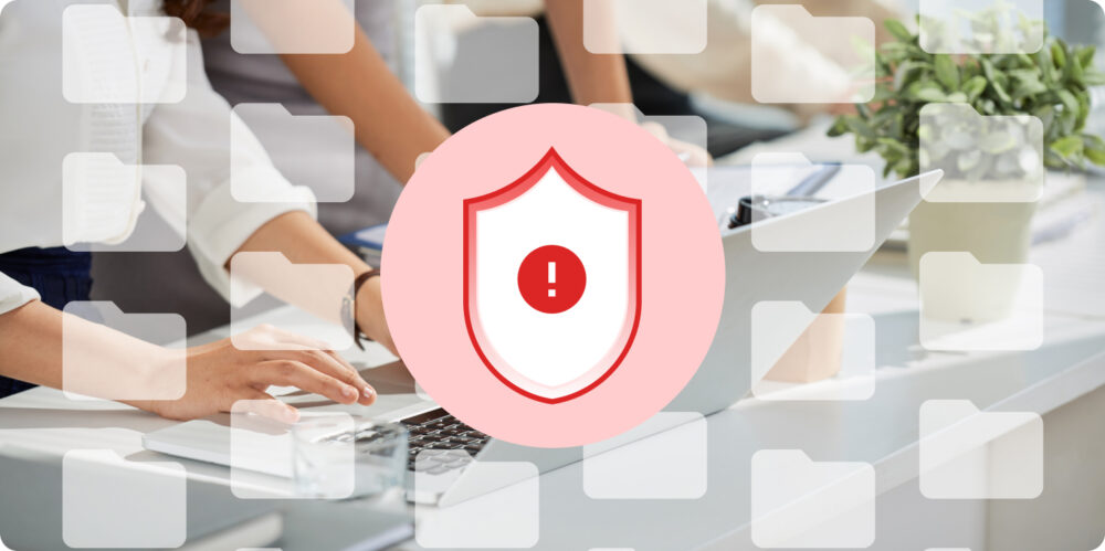 Personas trabajando en portátil con un escudo de advertencia y carpetas superpuestas; señales de la fragmentación de datos y riesgos de seguridad.