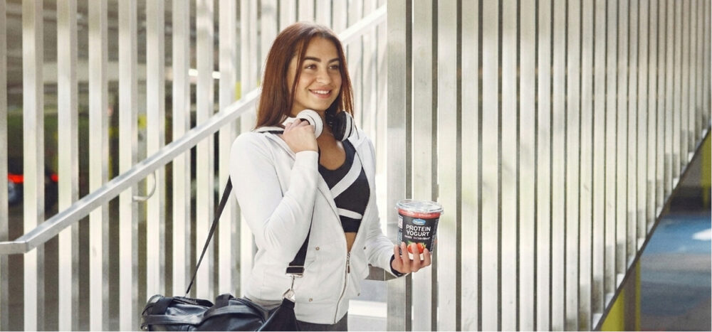 Mujer con bolsa de deporte sosteniendo un vaso de yogur de proteína en un entorno urbano.