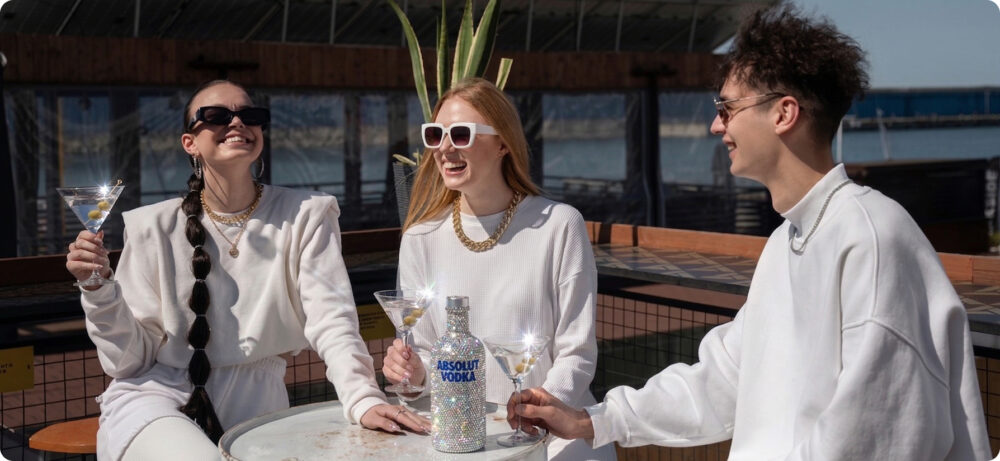 Três jovens sorrindo em um terraço enquanto seguram taças de coquetel ao lado de uma garrafa de vodka coberta de cristais, em uma cena de estilo bling-bling e consumo premium.