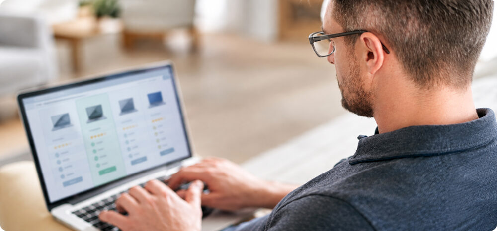 Análisis profesional y comparación de productos en tiendas online y e-commerce Vista desde el hombro de un profesional con gafas trabajando en su portátil mientras analiza y compara fichas de productos en un catálogo de comercio electrónico, evaluando precios, imágenes y características en un entorno de oficina en casa.